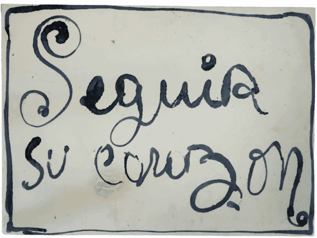 Scritta calligrafica nera “Seguía su corazón” su fondo bianco sporco, racchiusa da una cornice dipinta a mano, con la grande S iniziale avvolta in un ricciolo decorativo.