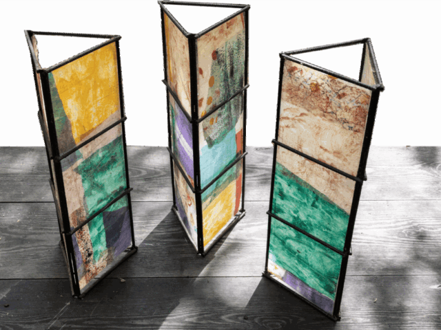 Tre strutture triangolari verticali in metallo nero, poggiate su assi di legno scuro, rivestite da pannelli di carta translucida a collage astratto con campiture gialle, verdi, viola e segni materici, simili a piccole vetrate colorate.
